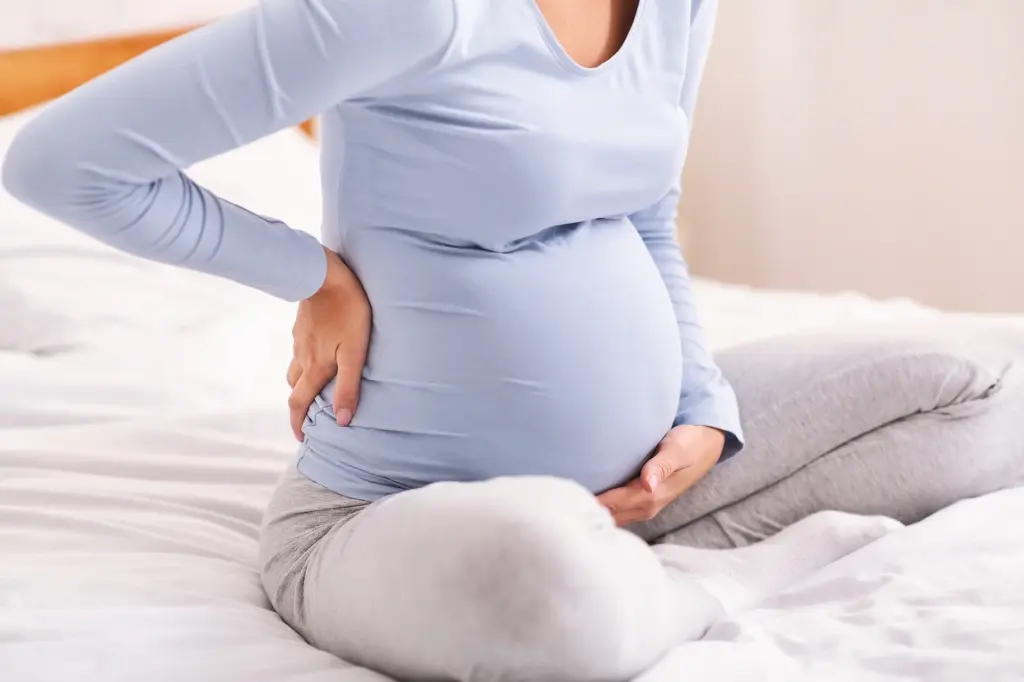 Schwangere Frau sitzt auf dem Bett, hält sich eine Hand an den unteren Rücken und stützt den Babybauch.