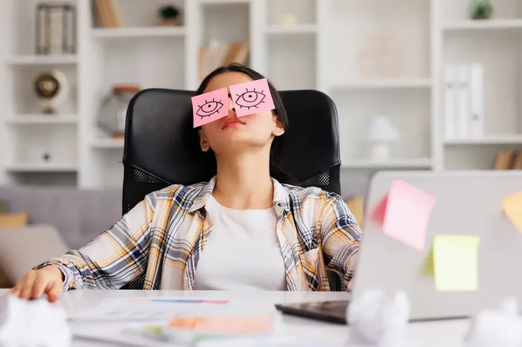 Erschöpfte Büroangestellte sitzt am Schreibtisch, lehnt im Bürostuhl zurück und hat gezeichnete Augen auf Haftnotizen über den geschlossenen Augen – Symbol für Müdigkeit, Stress und Erschöpfung am Arbeitsplatz.