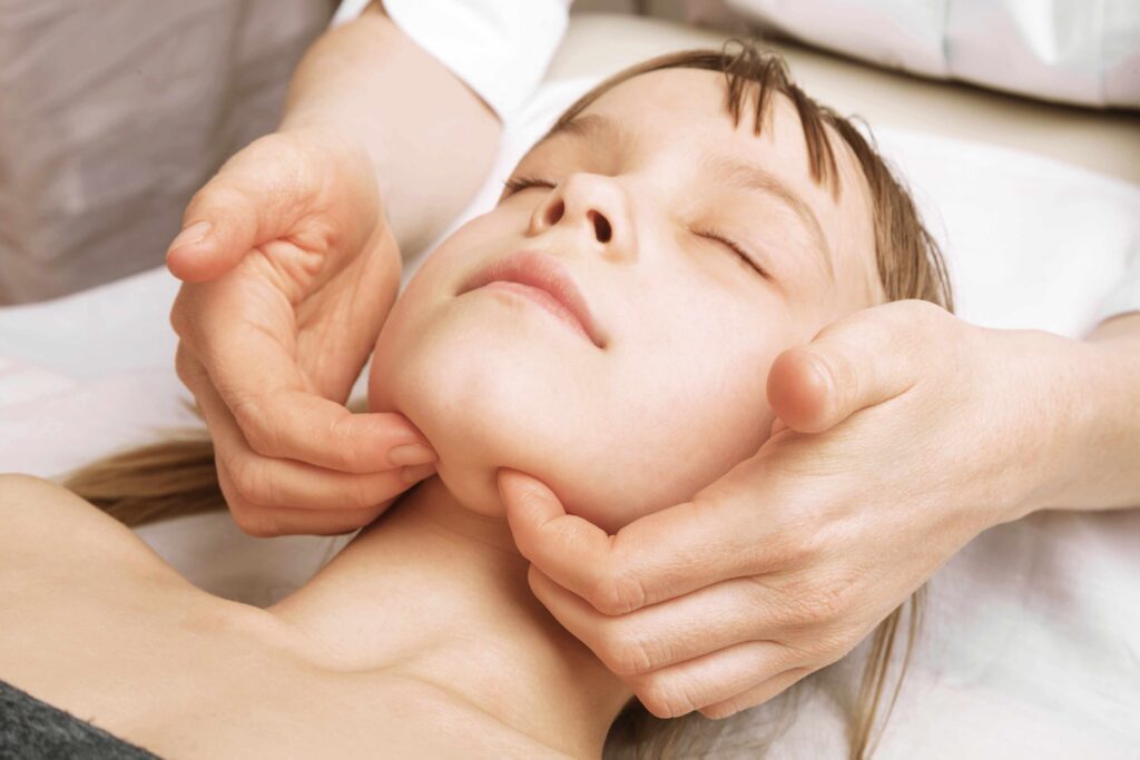 Kind liegt mit geschlossenen Augen, Therapeut berührt sanft Kinn und Nacken für eine sanfte Kieferbehandlung.